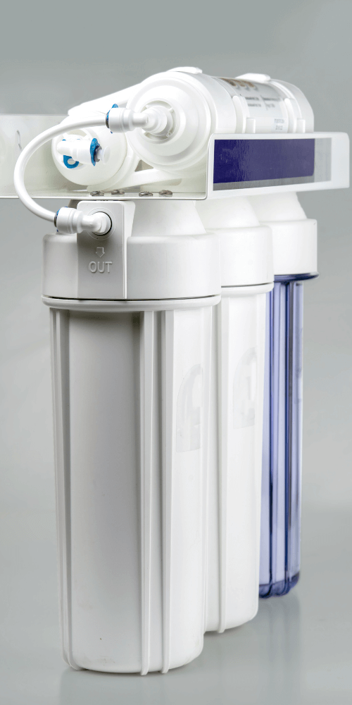 A close-up of a white water filtration system with three cylindrical canisters and a clear one, labeled "OUT" for filtered water.